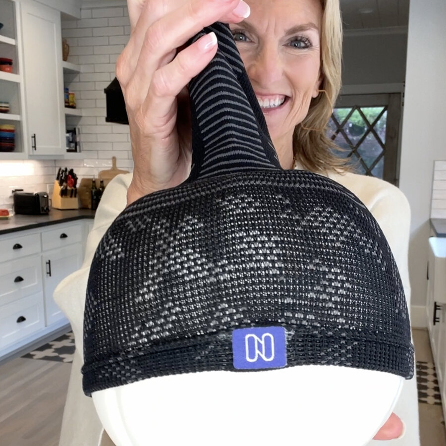 A woman in a kitchen holding one black sock up close to the camera, near enough to see the knit pattern and the woven cuff tag.
