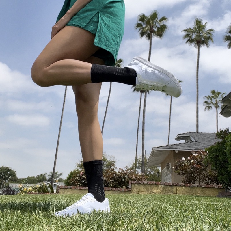 Black NanoSocks worn outdoors on grass under low-cut white sneakers, one leg raised mid-stride, with the ribbed top of each sock showing well above the shoe collar.